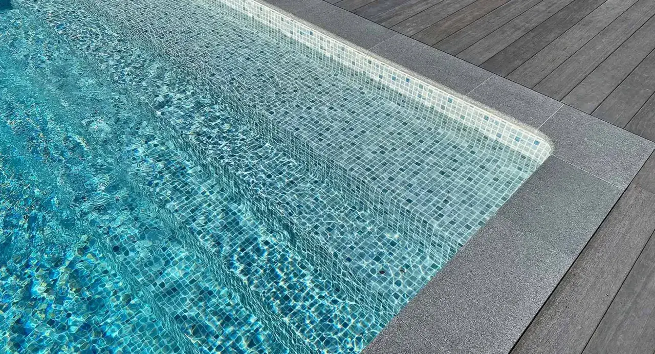 Trappsteg i en pool med blå mosaik, kantade av grå sten och trätrall. Sarg runt poolen ger en lyxig känsla.