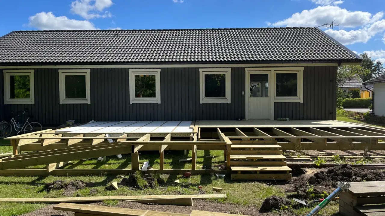 Bygga ut altan: Träkonstruktion för en ny altan tar form framför ett mörkt trähus. Solen skiner på en blå himmel med moln.