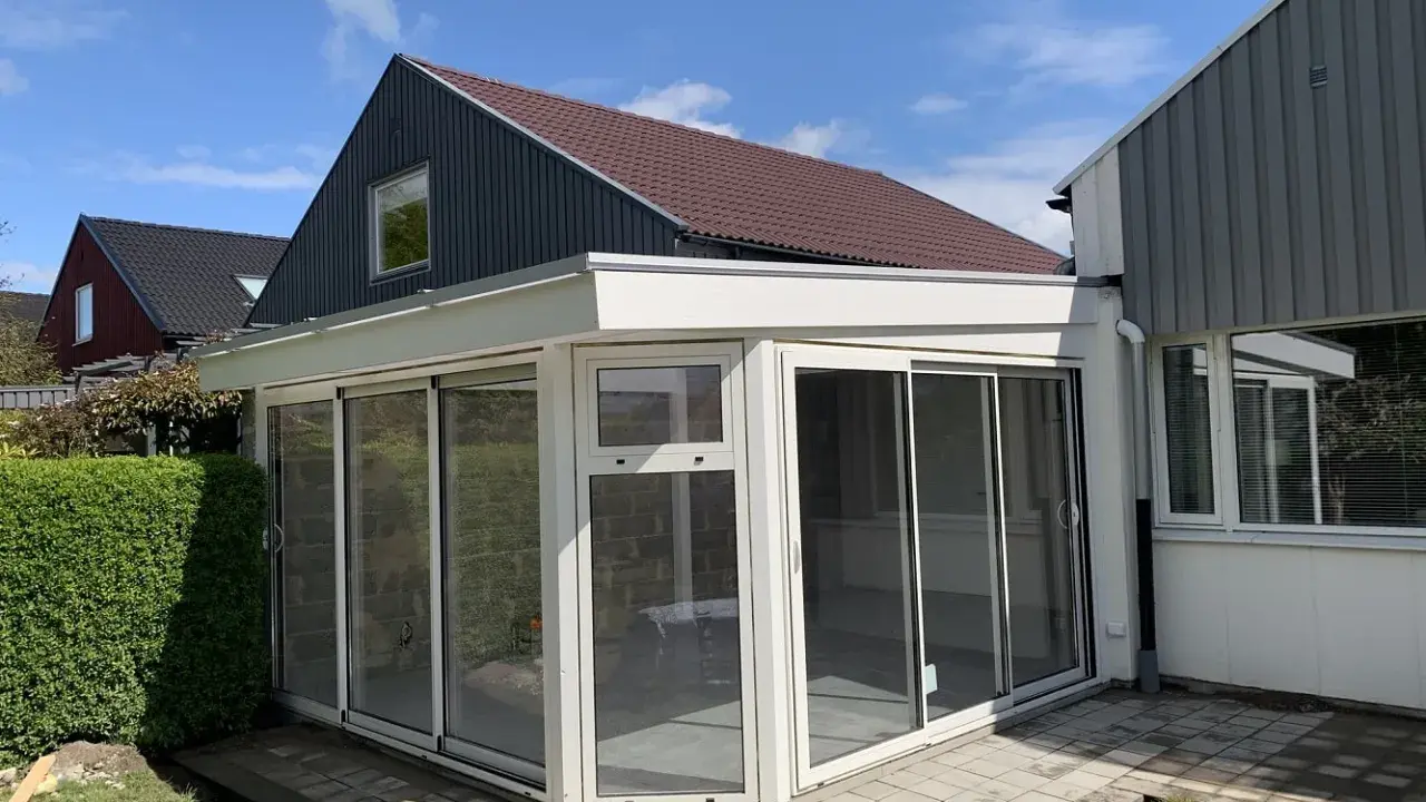 Ett modernt uterum med glaspartier och en vit takkonstruktion. Taklutningen på det angränsande huset ger en fin kontrast.