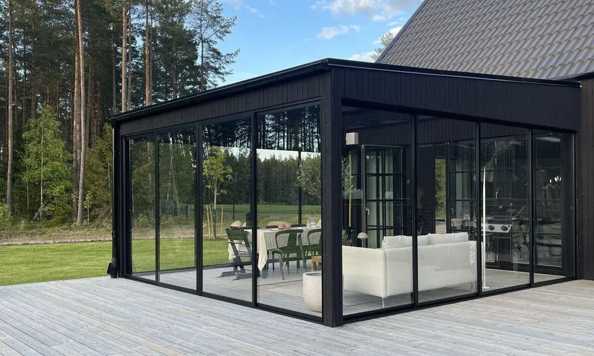 Svart glasveranda med matplats och soffgrupp, omgiven av skog. Trätrall som altan material ger en naturlig känsla.