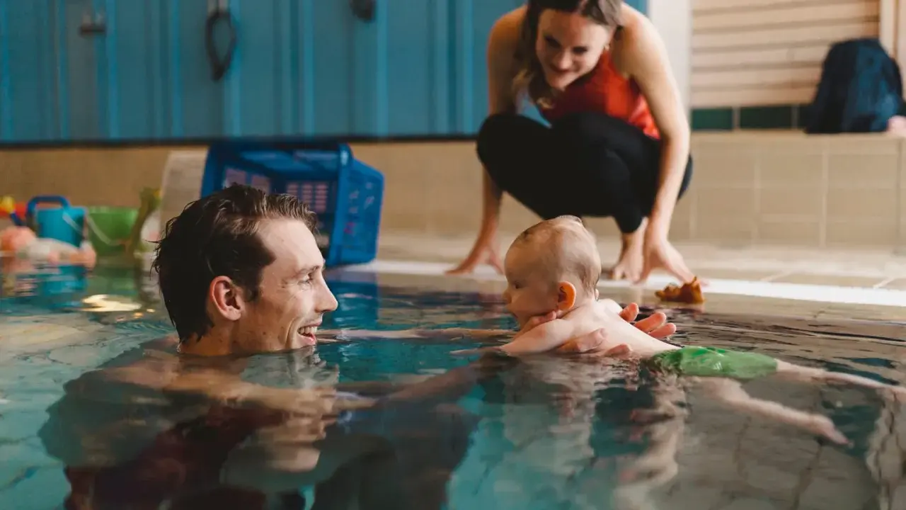 Glad pappa tittar på sitt bebis bada i poolen. En kvinna i bakgrunden ser också på.
