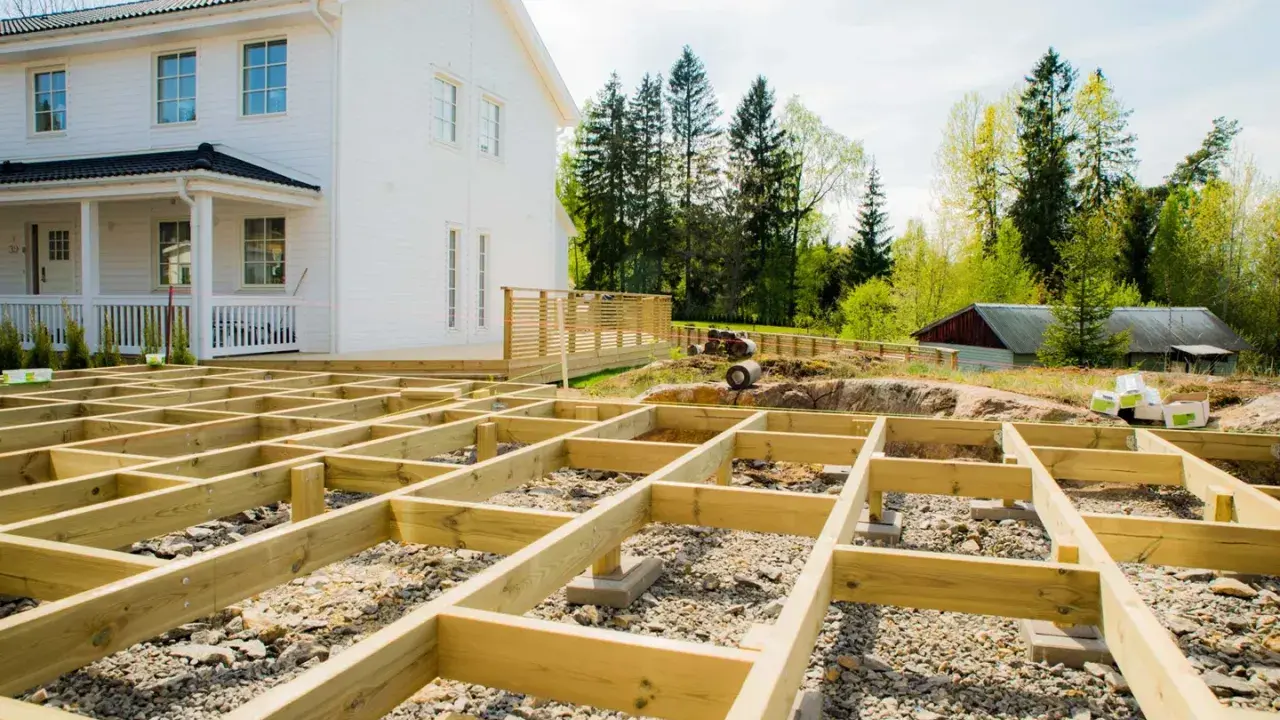 Grundarbete för att bygga altan på mark. Träkonstruktion på grus och sten framför ett vitt hus.