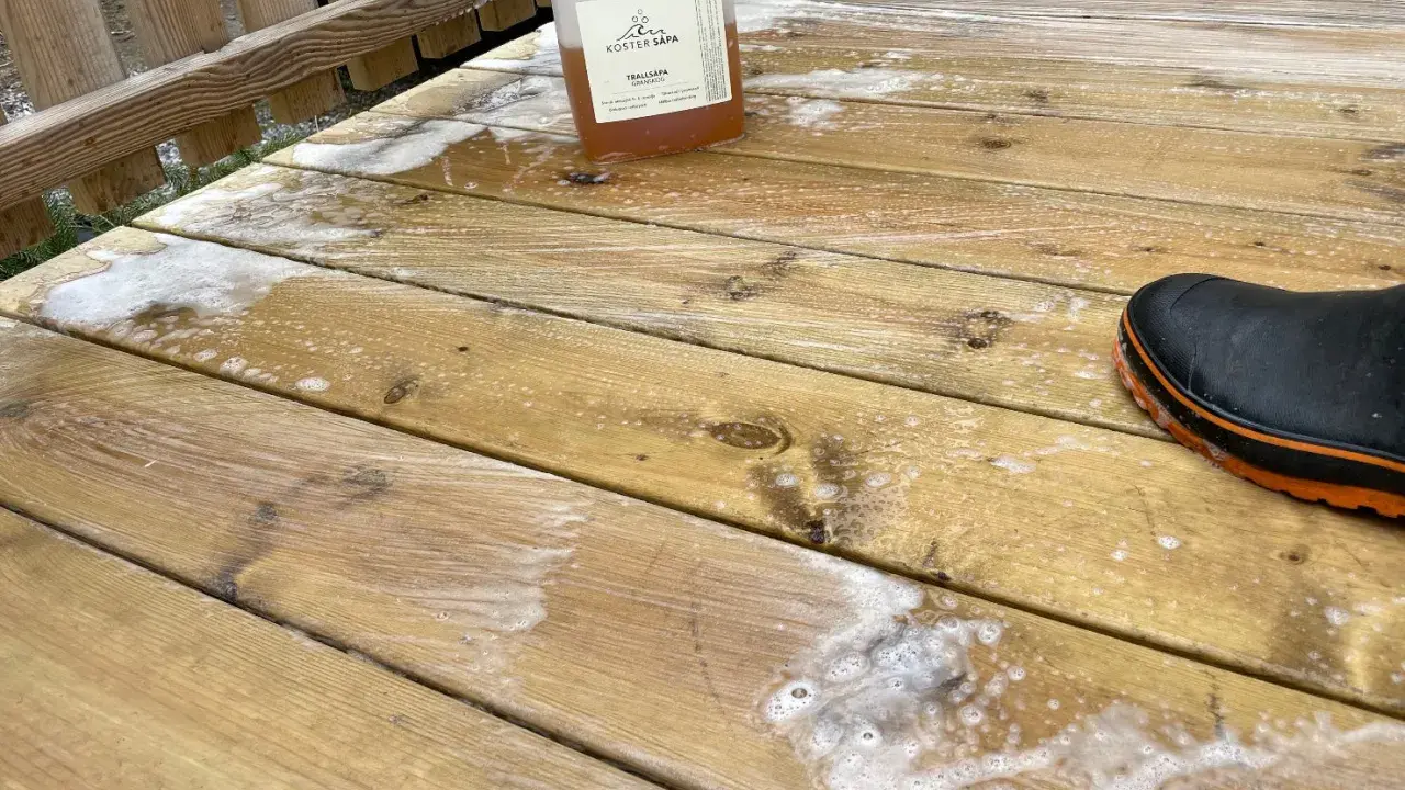 En flaska linoljesåpa står på en altan som håller på att rengöras. En stövel syns i bildens högra hörn.
