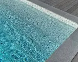 Trappsteg i en pool med blå mosaik, kantade av grå sten och trätrall. Sarg runt poolen ger en lyxig känsla.
