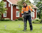 Man med orange jacka installerar skruvfundament för att **påla grund** till ett hus i trädgården.