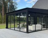 Svart glasveranda med matplats och soffgrupp, omgiven av skog. Trätrall som altan material ger en naturlig känsla.