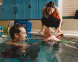 Glad pappa tittar på sitt bebis bada i poolen. En kvinna i bakgrunden ser också på.