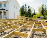 Grundarbete för att bygga altan på mark. Träkonstruktion på grus och sten framför ett vitt hus.