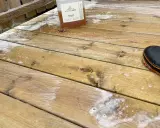 En flaska linoljesåpa står på en altan som håller på att rengöras. En stövel syns i bildens högra hörn.