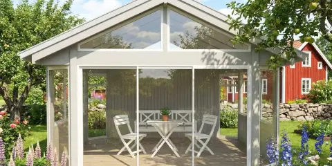 Ett fristående uterum på 25 kvm med glaspartier och skjutdörrar, möblerat med bord och stolar. Omgivet av en lummig trädgård med blommor och ett rött hus i bakgrunden.