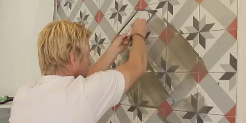 Man foga om kakel med en spackel. Mönstret på kaklet är geometriskt med stjärnor i grått, vitt och rött.