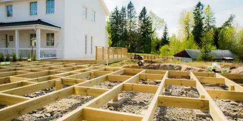 Grundarbete för att bygga altan på mark. Träkonstruktion på grus och sten framför ett vitt hus.