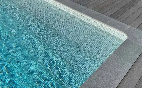 Trappsteg i en pool med blå mosaik, kantade av grå sten och trätrall. Sarg runt poolen ger en lyxig känsla.