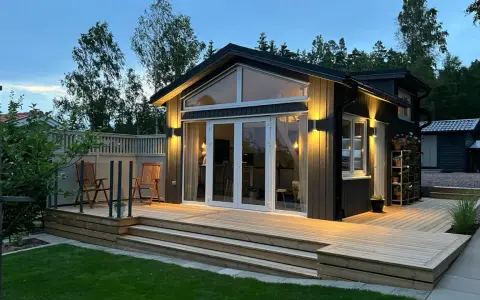Ett modernt hus med en stor, ljus träaltan som sträcker sig runt en del av huset. Två stolar står på altanen, redo för avkoppling.