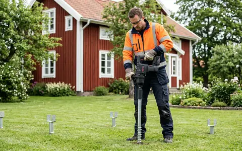 Man med orange jacka installerar skruvfundament för att **påla grund** till ett hus i trädgården.