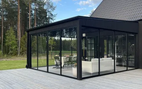 Svart glasveranda med matplats och soffgrupp, omgiven av skog. Trätrall som altan material ger en naturlig känsla.