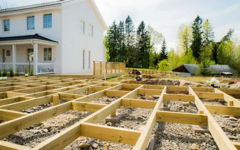 Grundarbete för att bygga altan på mark. Träkonstruktion på grus och sten framför ett vitt hus.