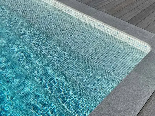 Trappsteg i en pool med blå mosaik, kantade av grå sten och trätrall. Sarg runt poolen ger en lyxig känsla.