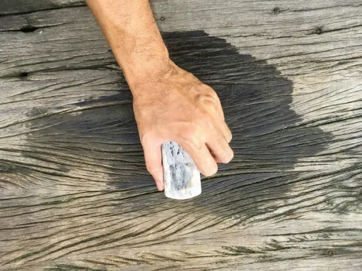 En hand håller en borste för att skura altanen. Träet är väderbitet och har en mörk, fuktig fläck där borsten nyss varit.