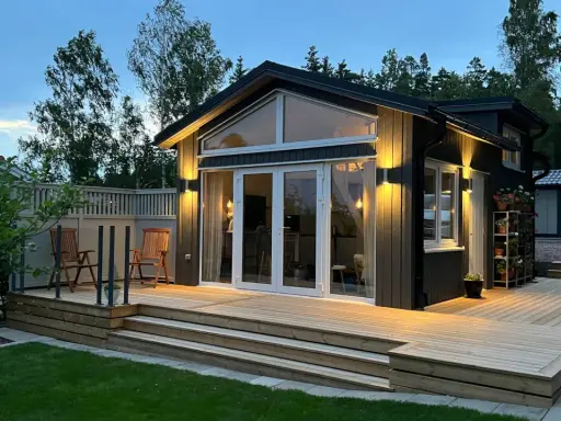 Ett modernt hus med en stor, ljus träaltan som sträcker sig runt en del av huset. Två stolar står på altanen, redo för avkoppling.