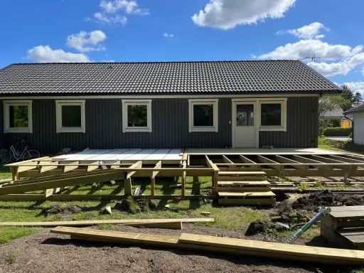 Bygga ut altan: Träkonstruktion för en ny altan tar form framför ett mörkt trähus. Solen skiner på en blå himmel med moln.