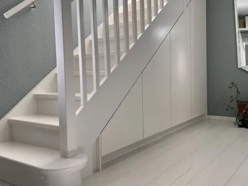 Vitt trappräcke och vita trappsteg leder uppåt. Utrymme under trappa har förvandlats till praktiska vita skåp.