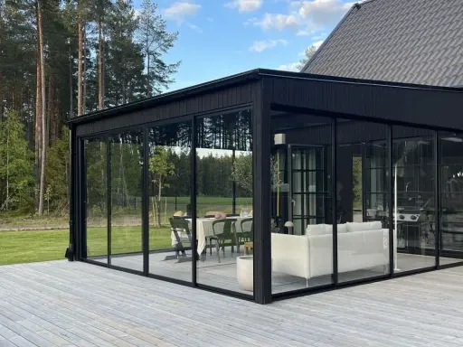 Svart glasveranda med matplats och soffgrupp, omgiven av skog. Trätrall som altan material ger en naturlig känsla.