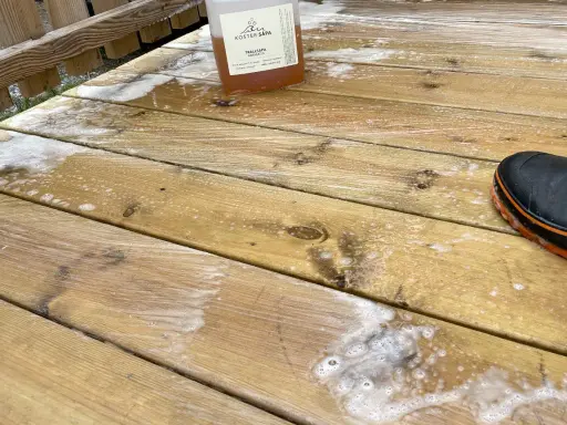 En flaska linoljesåpa står på en altan som håller på att rengöras. En stövel syns i bildens högra hörn.