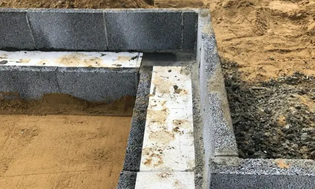 Grunden byggs med lecablock. Sand och grus ligger runt den pågående konstruktionen.