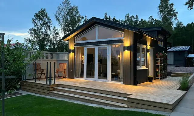 Ett modernt hus med en stor, ljus träaltan som sträcker sig runt en del av huset. Två stolar står på altanen, redo för avkoppling.