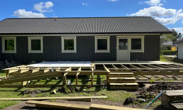 Bygga ut altan: Träkonstruktion för en ny altan tar form framför ett mörkt trähus. Solen skiner på en blå himmel med moln.