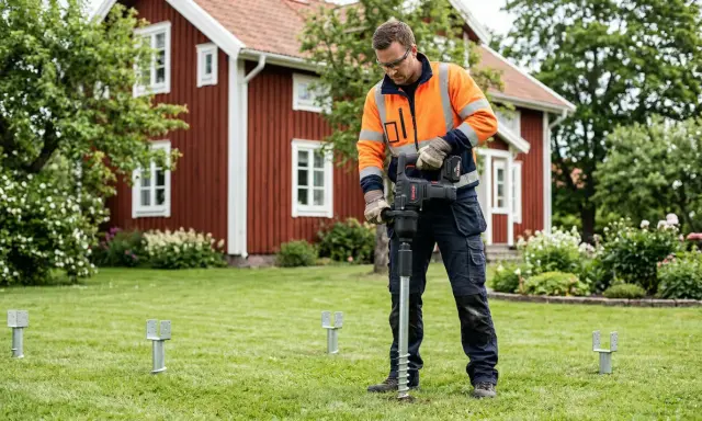 Man med orange jacka installerar skruvfundament för att **påla grund** till ett hus i trädgården.