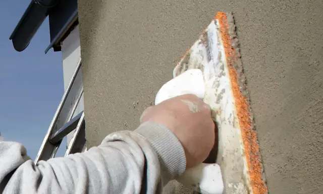 En hand med en murslev som arbetar med att putsa med murbruk på en fasad. En stege och stupränna syns i bakgrunden.