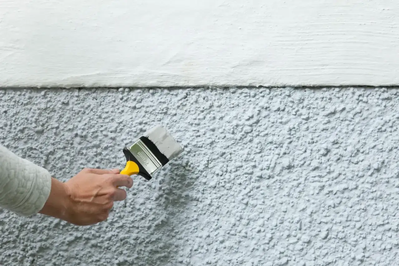 En hand målar en texturerad betongvägg utomhus med en pensel. En del av väggen är redan vitmålad.