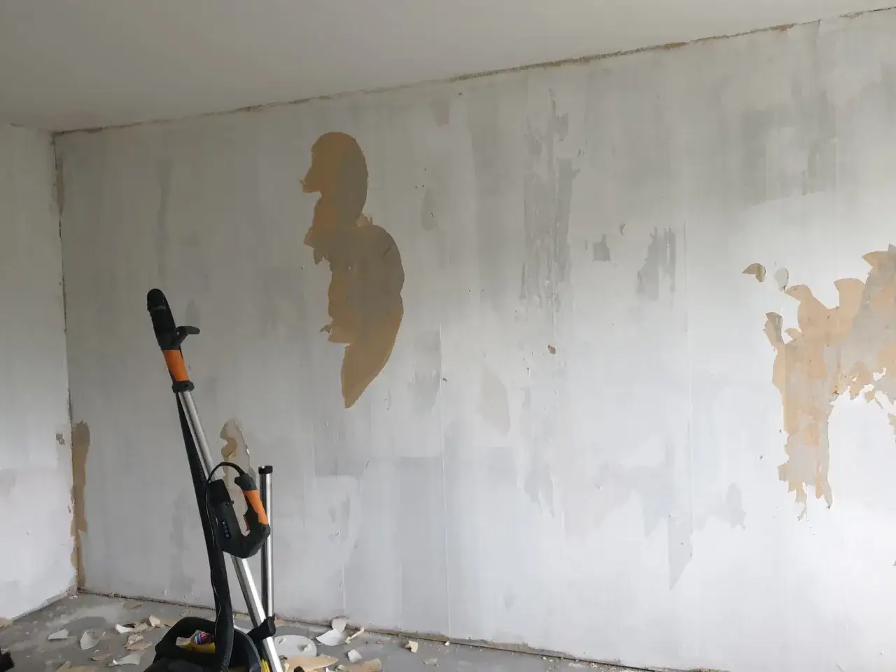 En v&auml;gg med gammal tapet som h&aring;ller p&aring; att tas bort, med sp&aring;r av bredspackla p&aring; tapet. En slipmaskin st&aring;r redo f&ouml;r att j&auml;mna till ytan.