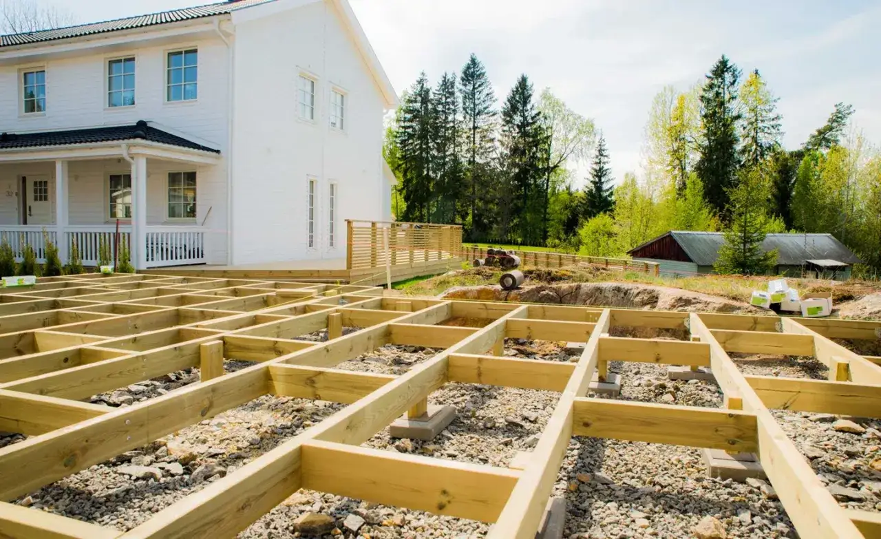Grundarbete f&ouml;r att bygga altan p&aring; mark. Tr&auml;konstruktion v&auml;xer fram framf&ouml;r ett vitt hus.