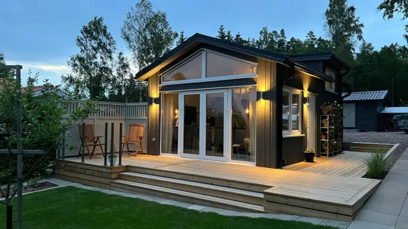 Ett modernt hus med en stor, ljus träaltan som sträcker sig runt en del av huset. Två stolar står på altanen, redo för avkoppling.